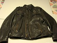 Front full view of a black Harley Davidson leather motorcycle jacket laid flat showing zippers, pockets, collar and side belts.