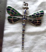 Dragonfly pendant laid flat on white fabric next to a ruler showing size approximately 3 inches long and 2.25 inches wide. Blue and black enamel on body, green enamel on wings with stained glass effect.