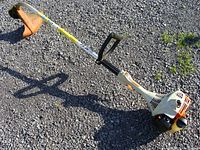 Full length view of Stihl gas weed eater showing white motor housing with orange parts and black handle, silver shaft, and orange cutting guard on gravel surface.