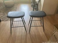 Photo of two black cushioned bar stools with aluminum legs on a hardwood floor, positioned side by side.