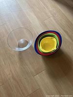 Photo showing one clear glass bowl next to a nested set of 10 colorful plastic salad bowls of varying sizes.
