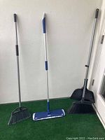 Photo of two brooms and a flat mop floor scrubber standing upright against a wall on green turf.