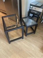 Three triangular-shaped black metal framed end tables with clear glass tops arranged on a light wood floor. The tables nest together to form one cohesive unit.