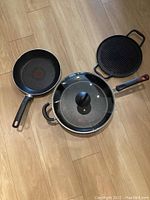 Photo of TFal non-stick frying pan, Tramontina cast iron skillet with glass lid, and another cast iron pan on wooden floor.