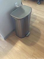 Tall aluminum garbage can with step pedal and closed lid, placed on wooden floor near wall outlet.