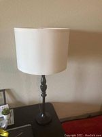 Table lamp with a white fabric cylindrical shade and dark aluminum turned stand placed on a side table against a beige wall.