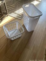 Photo shows 2 white plastic laundry baskets on a wooden floor with sunlight. One basket is tall and rectangular with side handles; the other is shallow and square with large ventilation holes and molded handles.