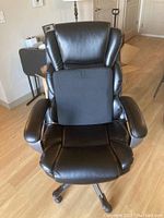 Front view of black padded swivel office chair with lumbar support pillow on wooden floor, visible minor wear and scuff marks.