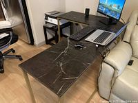 Full view of the L-shaped desk with metal frame and black marble-effect top, showing its placement beside a sofa and other office items.