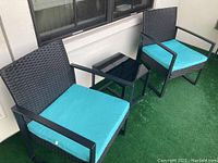 Two black woven rattan-style chairs with turquoise cushions placed on green artificial grass, accompanied by a black glass top table with metal frame.