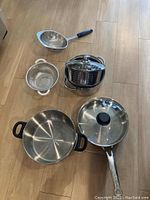 Photo showing 9 stainless steel steamer pots stacked, 2 metal strainers, a large pan with black handles, and a frying pan with glass lid, all placed on wooden floor.