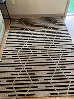 Photo showing a rectangular beige rug with black geometric linear and diamond patterns placed on light flooring near a wall and sliding glass door.