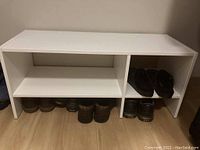 Image of white shoe storage shelf holding multiple pairs of shoes including boots and sneakers, placed on wooden floor against a beige wall.