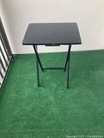 Black wood folding TV table with metal legs. The table is set up on green outdoor carpet next to a railing.