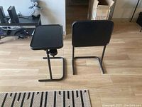 Photo showing two black folding metal TV tables with C-shaped bases and adjustable swivel tray tops positioned on a wood floor near a couch and TV.