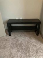 Photo of a black rectangular TV/entertainment stand with lower shelf against a light wall, positioned on beige carpet.