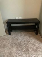 Black rectangular TV/entertainment stand with a lower shelf, placed against a beige wall and carpeted floor, showing overall condition and design