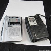 Two pocket radios placed on white paper, showing front view of both radios. Left is the silver Grundig Mini World 100 PE and right is the black Panasonic radio.