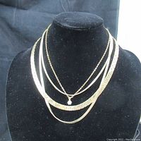 Three gold-tone chain necklaces on black velvet display including a small clear stone pendant, a wide flat chain, and a narrow box link chain