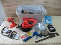 Wide shot of entire lot showing Black & Decker skillsaw, cordless flashlight, pipe wrench, hand saw blades, caulking gun, belts, small work light, black power adapter, and plastic storage bin with other tools and hardware inside.