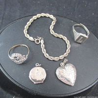 Photo showing all five jewelry items together on black stand: twisted rope chain bracelet, heart-shaped locket pendant, round locket pendant engraved 'G R', stone set silver ring, and signet ring with 'L' initial.