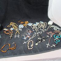 Wide shot showing multiple prayer beads, rosaries, metal crucifixes, and religious keychains laid out on black surface.