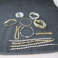 Full group of 11 bracelets on black cloth showing variety of styles and tones from silver to gold, beaded to chain links and bangles.