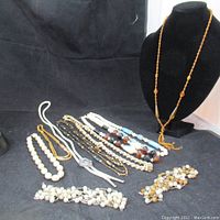 Image showing a collection of various necklaces, including faux pearl strands, beaded strands, and a bolo tie style necklace with turquoise centerpiece and silver metal tips, alongside earrings.