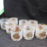 Photo of all seven matching glasses arranged on a black surface showing clear glass with gold sailing ship design and gold rim.