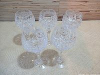 Five clear crystal glasses displayed on a beige surface, showing intricate cut patterns and stems.
