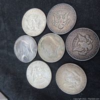 Seven US silver half dollar coins displayed on black fabric. Coins show obverse and reverse designs, varying degrees of wear and patina.