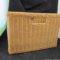 Side view of large rectangular wicker basket showing weave, handle, and structure against black backdrop.