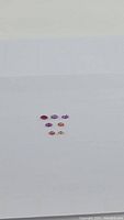 Seven marquis cut gemstones including one deep red ruby and six sapphires in shades of purple, pink, and orange laid out on white background.
