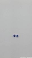 Pair of small, round-cut violet-blue tanzanite gemstones positioned side-by-side on a plain white background, showing size and color.
