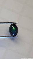 Oval cabochon cut black Ethiopian opal held with tweezers showing dark base color with green and blue color play.
