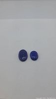 Two natural deep blue oval cabochon Lapis Lazuli gemstones on a white background, showing size difference and smooth polished finish.