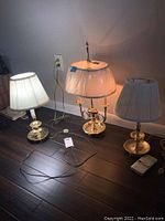 Three brass table lamps on dark wood floor near wall outlet. Two lamps have worn pleated shades and one has a shade still in plastic packaging. Electrical cords and plugs visible. Lamps around 25 inches tall.