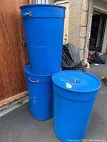 Three large blue plastic barrels stacked and placed next to a wooden surface. Two barrels are standing upright, one is stacked vertically on another.