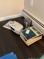 Photo of Bose Wave Radio/CD player, remote control on top, sheet music and music books stacked beside it, and CDs arranged vertically against the wall on dark hardwood floor.