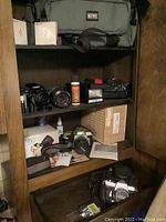 Photo shows cameras and accessories stored inside a wooden cabinet including Nikon N60 camera in original box, Minolta Maxxum 9000 camera body and zoom lens, carrying bags, flash, and cleaning supplies.