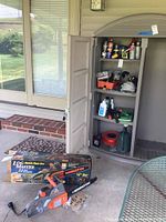 Remington electric chainsaw with orange and grey casing alongside its original branded box, placed next to a plastic shelving unit filled with workshop liquids and various tools.