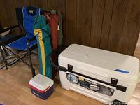 Full view showing one large white Igloo Glide wheeled cooler, one small blue and white Graco Rubbermaid lunch cooler, three canvas bag folding chairs in green, yellow, and red bags, and one blue folding chair with armrests and side table.