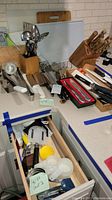Photo showing countertop full of various cutlery items including knives in blocks, cutting boards, cooking and serving utensils, white-handled knives, and boxed knives.