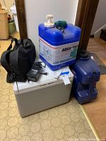 Full lot including Coleman cooler, two blue Aqua-Tainer water jugs, black bocce ball bag with power adapter on top of cooler