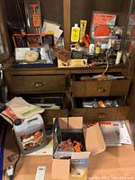 Wide shot of a wooden cabinet with drawers containing various firearm accessories, cleaning supplies, boxes, bottles, and other items. Cardboard boxes and containers on floor including a Beeman pellet trap box.