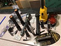 Overview of eleven flashlights of various sizes including a large rechargeable yellow DeWalt spotlight, black metal and plastic flashlights, clip-on light, protective goggles, and tape measure level. Items shown on a workbench surface.