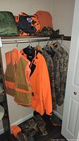 Closet rod with blaze orange safety mesh vest, orange hooded jacket, camouflage jackets; top shelf with green quilted item and orange camouflage bag