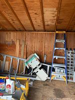 Two A-frame Werner ladders leaning against wood wall in shed, including one 6 ft and one 10 ft ladder, surrounded by other storage items.