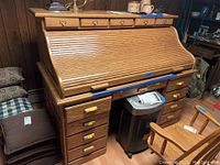 Full front view of oak roll top desk closed with visible multiple drawers and brass handles, chair beside the desk.
