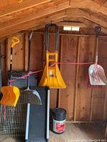 View of shovels hanging inside wooden shed including yellow The Bully shovel and plastic snow shovels with bucket of ice melt below.